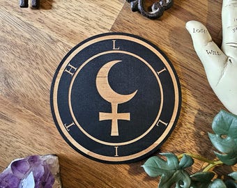 May include: A round, black wooden coaster with a light brown border. The coaster features a crescent moon and cross symbol, with the letters "L", "I", "H", and "T" around the edge. An antique key and amethyst crystal are also visible.
