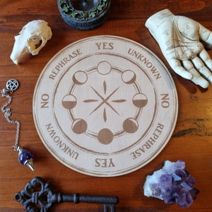 May include: A wooden Ouija board with a moon phase design. The board has the words "Yes", "No", "Rephrase", and "Unknown" around the edge.