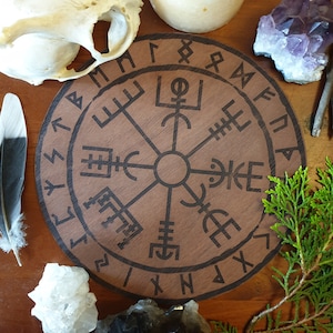 May include: A brown wooden circle with a black compass design and runes around the edge. The compass has four points with symbols at each point. The runes are in a circle around the compass. The circle is on a wooden surface.