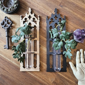 May include: Two wall-mounted gothic-style wooden vases, one natural and one black, each holding a glass test tube vase with green leafy stems. The vases are on a wooden surface, with a key and other decorative items.