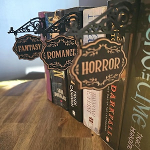 以下が含まれることがあります： 「FANTASY」「ROMANCE」「HORROR」と書かれた装飾的な看板が付いた黒い金属製のブックエンド。看板はヴィンテージスタイルで、本を並べています。