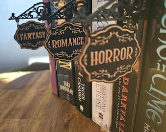 May include: Black metal bookends with decorative scrollwork and hanging signs that read "FANTASY", "ROMANCE", and "HORROR". The signs are in a vintage style, and the bookends are holding a row of books.