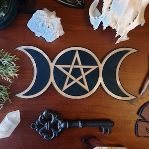 May include: A wooden triple moon and pentacle wall hanging with a black and natural wood finish. The design features three crescent moons surrounding a central pentagram. A decorative key and crystals are also visible.