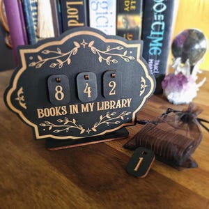 Puede incluir: Un contador de biblioteca de madera negra con el texto "BOOKS IN MY LIBRARY" y los números 842. El contador tiene un borde dorado decorativo y detalles florales. Una pequeña ficha de madera con el número 1 está delante.