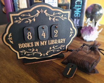 May include: A black wooden library counter with the text "BOOKS IN MY LIBRARY" and the numbers 842 displayed. The counter has a decorative gold border and floral accents. A small wooden token with the number 1 is in front of the counter.