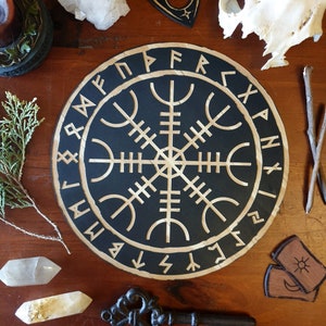Puede incluir: Un letrero redondo de madera negra con un símbolo Vegvisir blanco en el centro. El símbolo está rodeado por un círculo de runes. El letrero está sobre una superficie de madera.