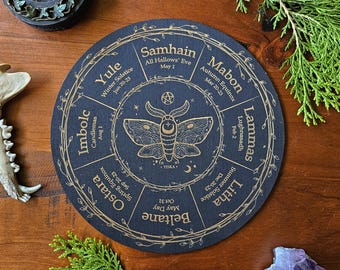 May include: A black circular wheel with gold text and illustrations depicting the eight sabbats of the Wheel of the Year. The wheel is divided into eight sections, each representing a different sabbat. The sabbats are: Yule, Imbolc, Ostara, Beltane, Litha, Lammas, Mabon, and Samhain. The wheel also features a stylized moth illustration in the center.