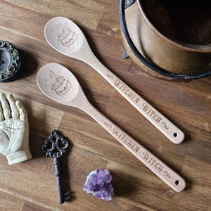 May include: Two wooden spoons with the text "KITCHEN WITCH" engraved on the handles. Each spoon features an engraved cauldron design on the bowl. The spoons are light brown and rest on a wooden surface.