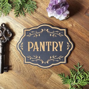 Può includere: Un cartello per dispensa in legno nero e marrone con la scritta "PANTRY" in grandi lettere dorate. Il cartello ha un bordo decorativo con accenti floreali. Il cartello è su una superficie di legno con una chiave, ametista e vegetazione.