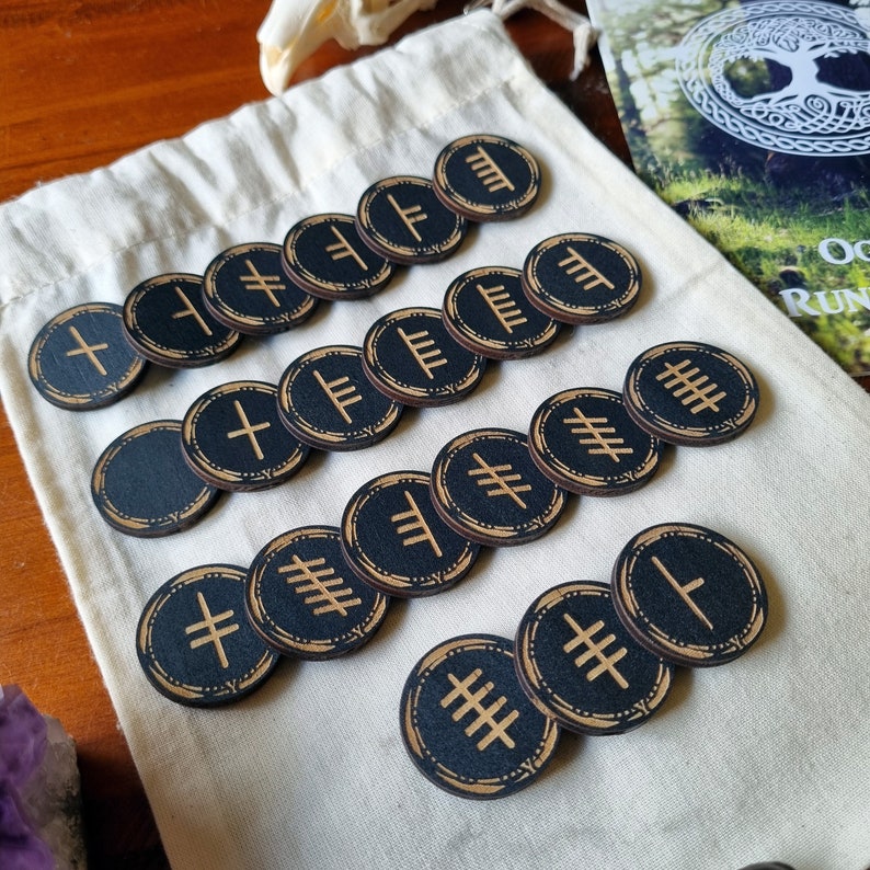 May include: A set of 18 black wooden runes with gold symbols. The runes are arranged in rows on a white fabric surface.