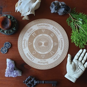 May include: A wooden wheel with the names of the eight sabbats of the Wheel of the Year, including Yule, Imbolc, Ostara, Beltane, Litha, Lammas, Mabon, and Samhain. The wheel is decorated with a Celtic knot design and a tree of life in the center.