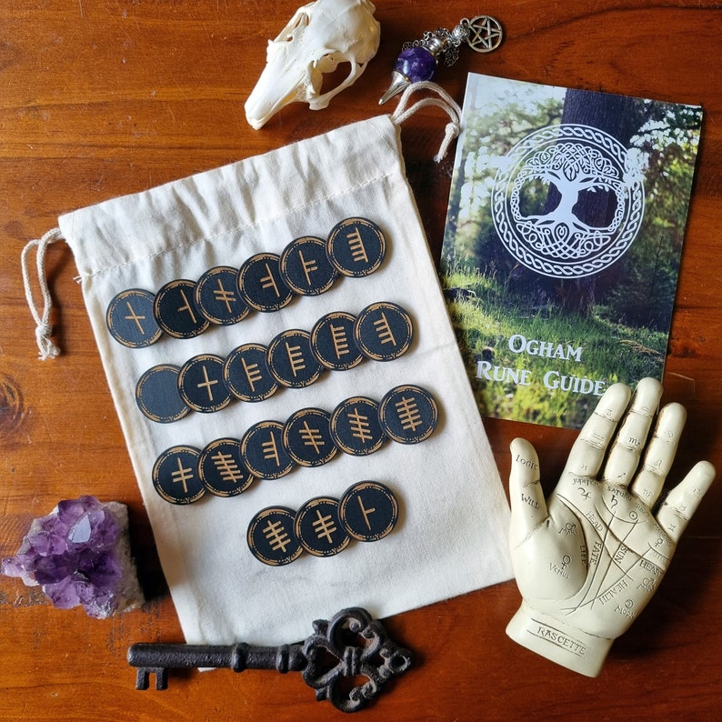 May include: A set of 15 black and gold Ogham runes, with a white cloth bag, a book titled "Ogham Rune Guide", and a palmistry hand. The runes are circular and feature a different symbol on each one.