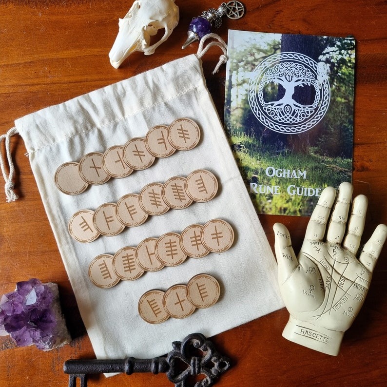 May include: A set of wooden Ogham runes in a white cloth bag. The runes are circular and have different symbols carved into them. The bag is sitting on a wooden table with a book titled "Ogham Rune Guide" and a hand with lines for palmistry.