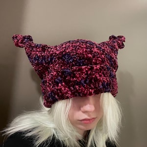 May include: A handmade, textured hat in shades of burgundy and purple. The hat features pointed ear-like protrusions at the top. The hat is made of a soft, plush material, perfect for adding a unique touch to any outfit.