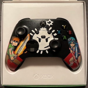 May include: Black Xbox controller with custom artwork featuring cartoon characters and a white sun-like design. The controller has colored buttons and directional controls. The Xbox logo is visible at the bottom.