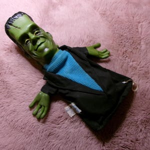 May include: A Frankenstein monster hand puppet featuring a moulded green plastic head with black hair and a detailed facial expression. The puppet wears a blue ribbed shirt under a black jacket, with green plastic hands attached to the sleeves.