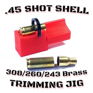 Pode incluir: Um gabarito de corte vermelho .45 Shot Shell com uma alça preta, segurando um estojo de latão. Abaixo, o texto diz "308/260/243 Brass Trimming Jig". Dois estojos de latão também são visíveis.