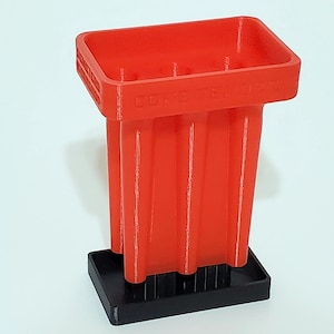 May include: A red plastic cone tender with the text "CONE TENDER" on the side. The cone tender has a rectangular top and several vertical columns. It sits on a black rectangular base.