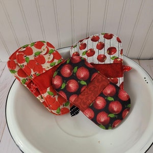 May include: Three red and white apple-themed oven mitts. The mitts are made of fabric and have a quilted design. They are sitting in a white enamel bowl.