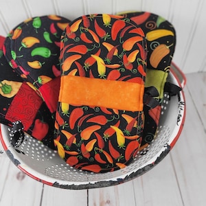 May include: A collection of oven mitts and pot holders with a chili pepper pattern in a white and red colander. The mitts and holders feature a black background with red, orange, and yellow chili peppers. An orange band is sewn on the mitt.