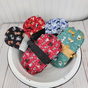 May include: A collection of five fabric pot holders with various animal prints, including cats and dogs, in red, black, blue, and teal colors. Each pot holder features a contrasting fabric band across the center and is arranged inside a white bowl.