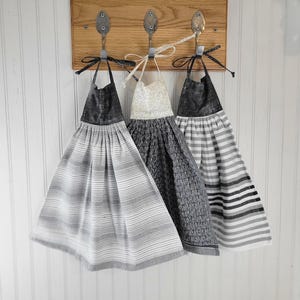 May include: Three decorative kitchen towels hanging on a wooden board with spoon hooks. The towels feature various patterns in shades of gray and white, including stripes and textured designs. The board has a sign that reads "IMAGINE-CREATE-INSPIRE OPEN 24 HOURS."