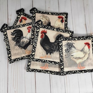 May include: A set of four square fabric pot holders featuring illustrations of roosters in various colors including black, white, and grey. Each pot holder is finished with a black fabric border decorated with a small white floral pattern.