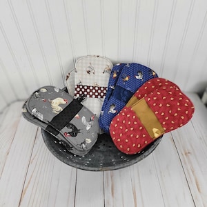May include: A collection of oven mitts in a metal colander. The mitts feature various patterns, including chickens on gray, polka dots, and red with small white dots. Each mitt has a black or brown band.