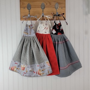 May include: Three hanging kitchen towels with fabric tops and textured bottoms. The left towel has a gray chevron pattern and kitchen tool print. The center towel is solid red with a white kitchen tool top. The right towel is gray with a black top.