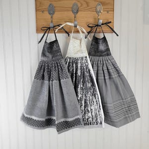 May include: Three decorative kitchen towels hanging on a wooden board with spoon hooks. The towels feature various gray patterns, including geometric and abstract designs. A sign above reads "IMAGINE-CREATE-INSPIRE OPEN 24 HOURS."