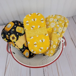 May include: Three fabric oven mitts with distinct patterns: one black with sunflowers and honey jars, one yellow with a bee print, and one yellow with a white daisy and bee print. The mitts are arranged inside a white metal colander with a red rim.