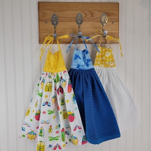 May include: Three decorative kitchen towels hanging on a wooden board. The towels feature colorful patterns: one with bugs, one with butterflies, and one with daisies. The board has a metal sign that says "Sewing Imagine-Create-Inspire Open 24 Hours."