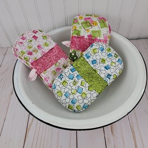May include: Three colorful oven mitts with geometric patterns in pink, green, and blue. The mitts are made of fabric and have a quilted design. They are sitting in a white bowl with a black rim.