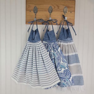May include: Three hanging kitchen towels with blue denim-style tops and tie closures. The first towel features horizontal gray and white stripes, the second has a blue and white paisley pattern, and the third displays wide blue and gray horizontal stripes.