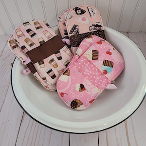May include: Four pink and brown oven mitts with different patterns. One mitt has a coffee cup design, another has a cupcake design, and the other two have a teacup design. The mitts are in a white bowl.