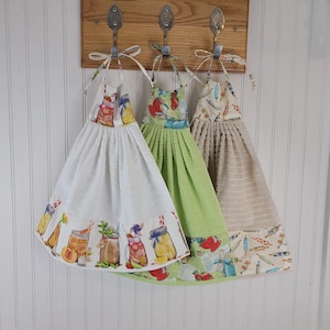 May include: Three hanging kitchen towels. The first is white with a colorful mason jar design. The second is green with a red and green pattern. The third is tan with a blue and tan pattern. All are hanging on a wooden board.