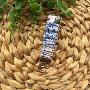 May include: A fabric wristlet keychain with a brown leather-like tab and a silver-colored key ring. The fabric is white with blue floral and abstract patterns, and red and green accents.