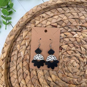 May include: A pair of dangle earrings featuring a layered design with a small black diamond-shaped top, a white semi-circle with black speckles in the middle, and a black scalloped fan-shaped base, all attached to silver-toned metal earring hooks.
