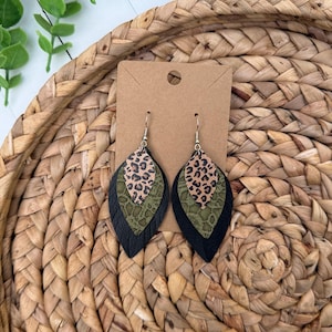 May include: A pair of layered leaf-shaped earrings featuring three distinct textures: a top layer with a tan and black leopard print, a middle layer in olive green with a textured lace pattern, and a bottom layer in solid black, all attached to silver hooks.