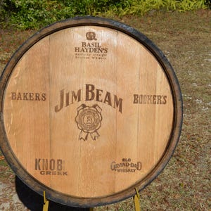 May include: A wooden barrel head with the Jim Beam logo and the names of other bourbon brands. The barrel head is made of light brown wood with dark brown trim. The text includes "Bakers", "Bookers", "Knob Creek", and "Old Grand-Dad Whiskey".