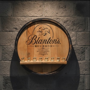 May include: A circular wooden barrel head display rack featuring the black printed text Blanton's BOURBON THE ORIGINAL SINGLE BARREL BOURBON and a horse and jockey logo. A horizontal shelf holds eight small gold-colored horse and jockey figurines.