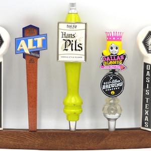 May include: A wooden beer tap handle rack with eight different beer tap handles. The handles are made of wood, metal, and plastic and feature various logos and designs. The rack is made of dark wood and has a curved shape.