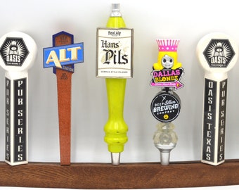 Op de afbeelding: Een houten biertapkraanrek met acht verschillende biertapkranen. De kranen zijn gemaakt van hout, metaal en plastic en hebben verschillende logo's en ontwerpen. Het rek is gemaakt van donker hout en heeft een gebogen vorm.
