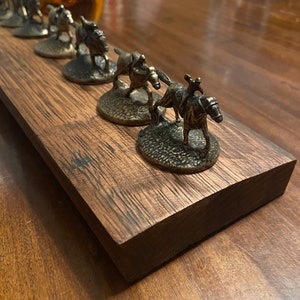 May include: A wooden board with six miniature bronze horse and jockey figurines. The figurines are arranged in a line, with the horses facing forward. The board is stained a dark brown color and has a rustic appearance.