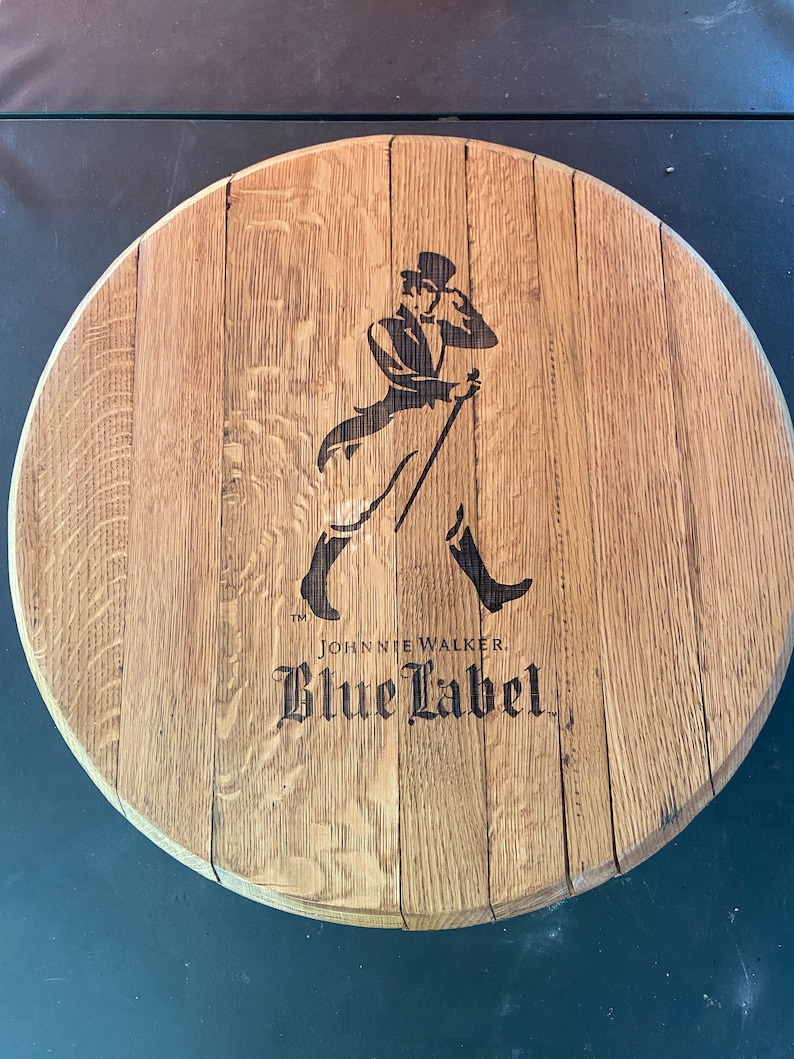 Pu&ograve; includere: Un coperchio di botte di legno rotondo con un'immagine in bianco e nero di un uomo con un cappello a cilindro e un bastone. Il testo "Johnnie Walker Blue Label" &egrave; stampato sotto l'immagine.