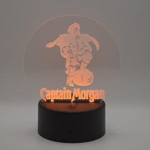 May include: A 3D printed night light with a clear acrylic base and a round, translucent acrylic top. The top features a silhouette of a cartoon pirate holding a bottle of rum. The text "Captain Morgan" is printed below the silhouette.