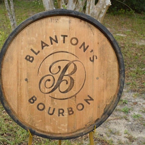 May include: Wooden barrel head with the words "Blanton's Bourbon" in a circular design. The letter "B" is stylized in the center. The barrel head is brown with a black rim and is displayed outdoors.