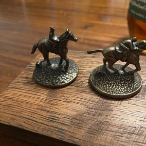 May include: Two miniature bronze horse and rider figurines on a wooden base. The horses are in a racing pose with jockeys on their backs. The base is made of dark wood with a rough texture.