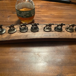 May include: A wooden board with nine miniature bronze horse figurines, each with a jockey, arranged in a line. The figurines are on a wooden board with a dark brown finish.