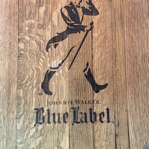 Pu&ograve; includere: Una botte di legno con un'immagine in bianco e nero di un uomo con un cappello a cilindro e un bastone. Il testo "Johnnie Walker Blue Label" &egrave; stampato sotto l'immagine.